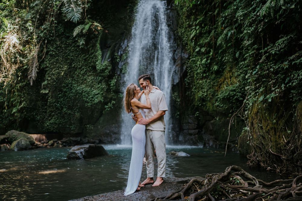 A couple poses together near a tropical waterfall with fallen logs in the foreground.