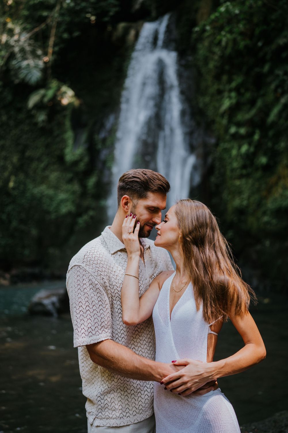 Two people in white outfits share a tender moment against a backdrop of tropical foliage.