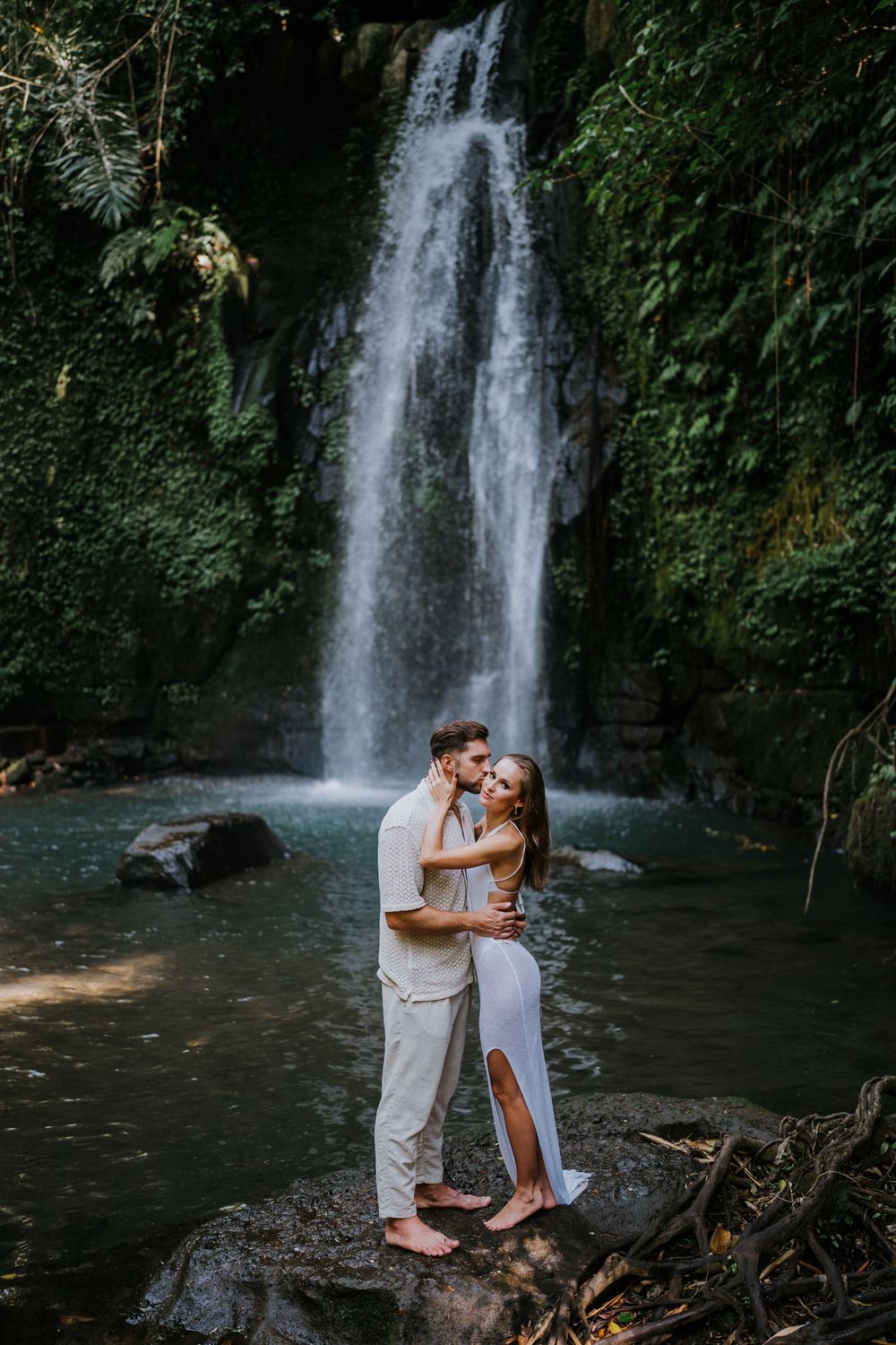 A couple shares an intimate moment in front of a cascading waterfall surrounded by greenery.