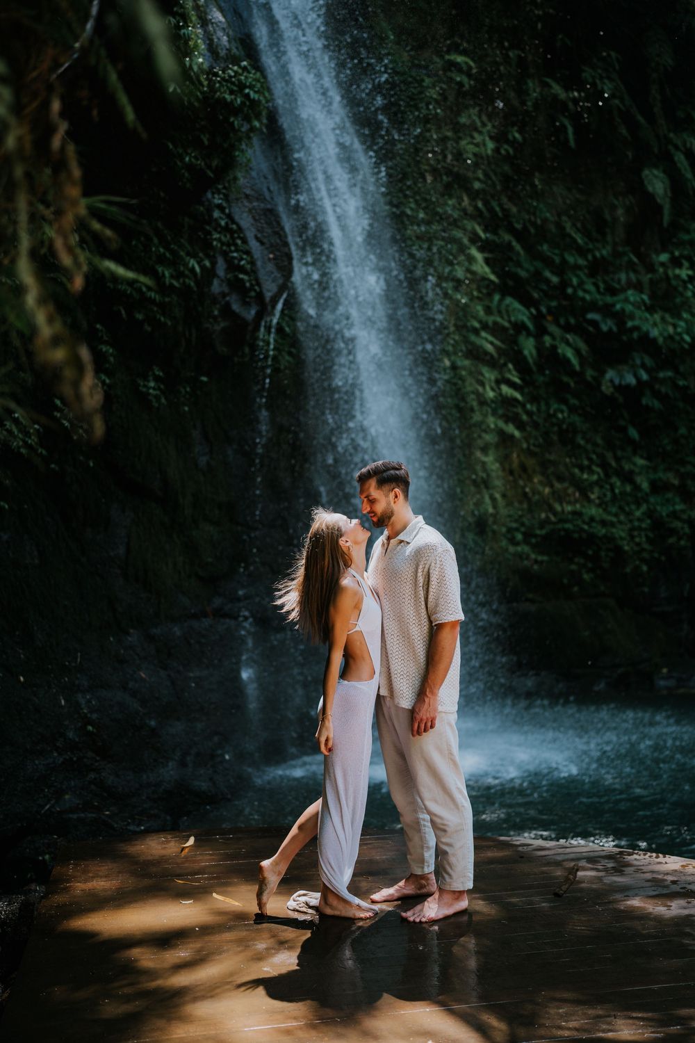 A couple in white outfits poses together by a scenic waterfall in a tropical setting.