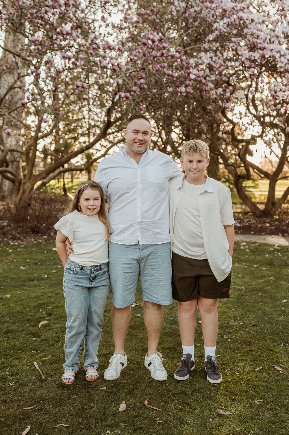 People wearing light colored shirts and denim stand together on grass near blooming trees at sunset.