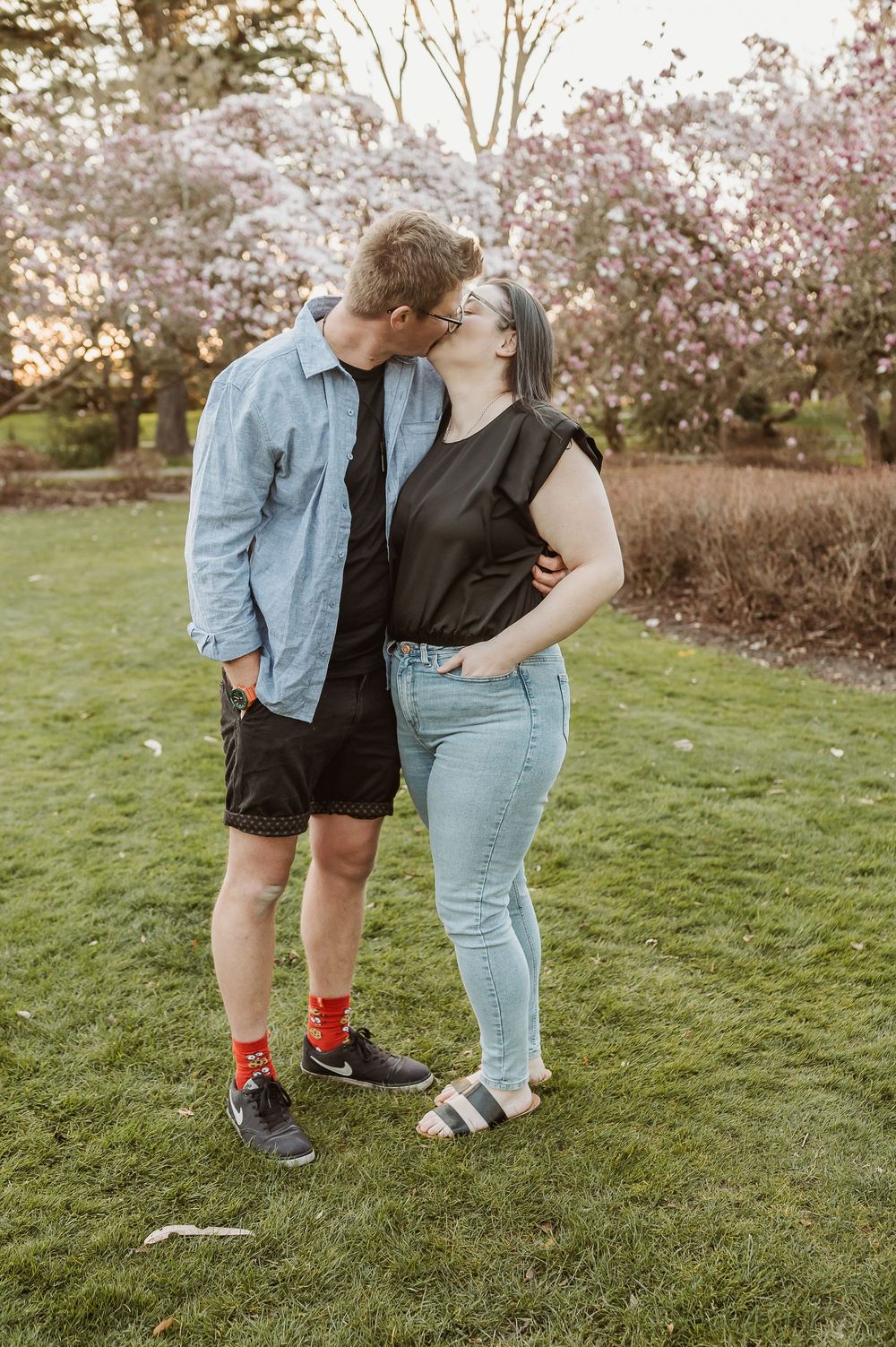 Two people share an intimate moment in a garden with blooming cherry blossom trees in the background during sunset.