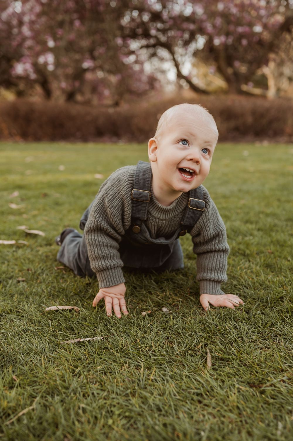 A young child in a gray sweater crawls on green grass while smiling and looking up playfully.