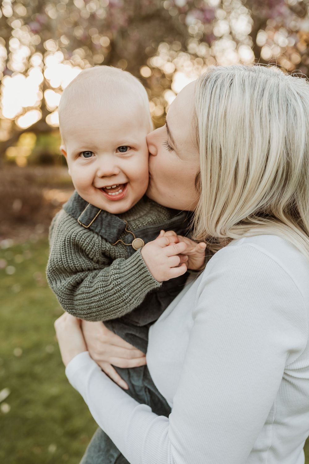 A tender moment captured outdoors with a joyful baby in a green sweater being kissed during golden hour.