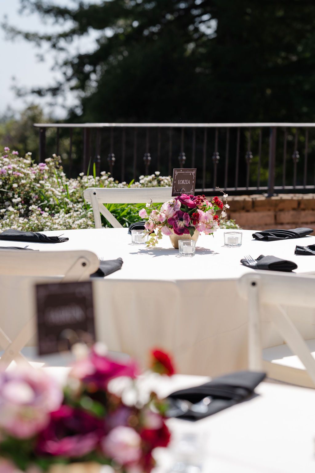 An elegant outdoor table setting with white tablecloths and pink floral centerpieces against a blurred forest background.