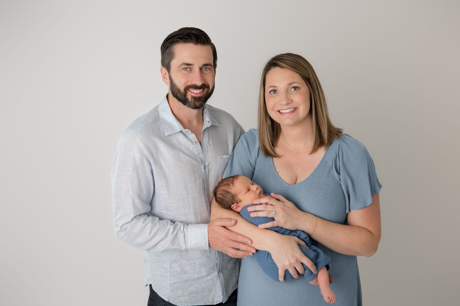 A couple embraces while holding their newborn baby in a casual indoor photoshoot setting.