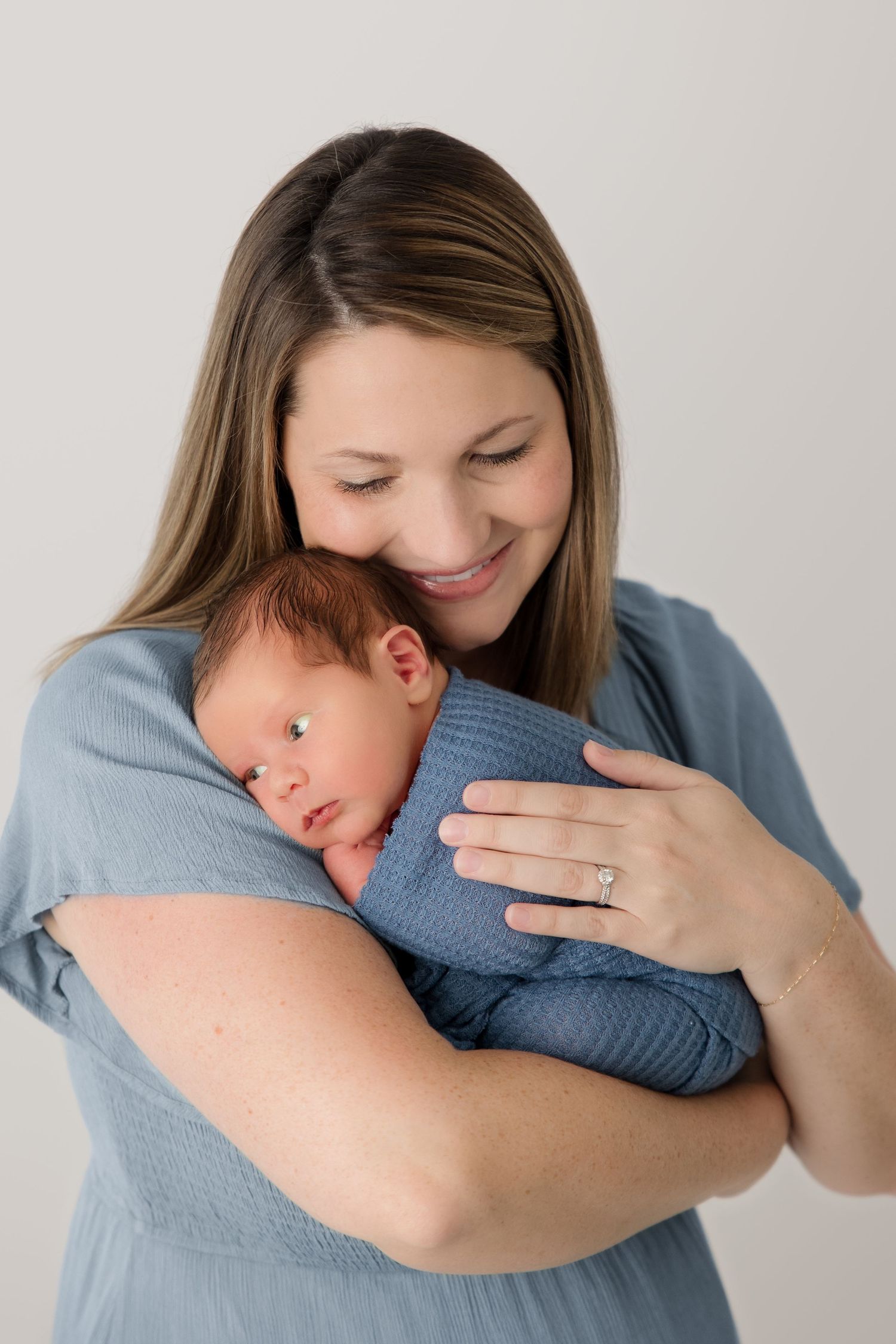 A woman in a blue shirt tenderly embraces a baby wearing a blue outfit against a light background.