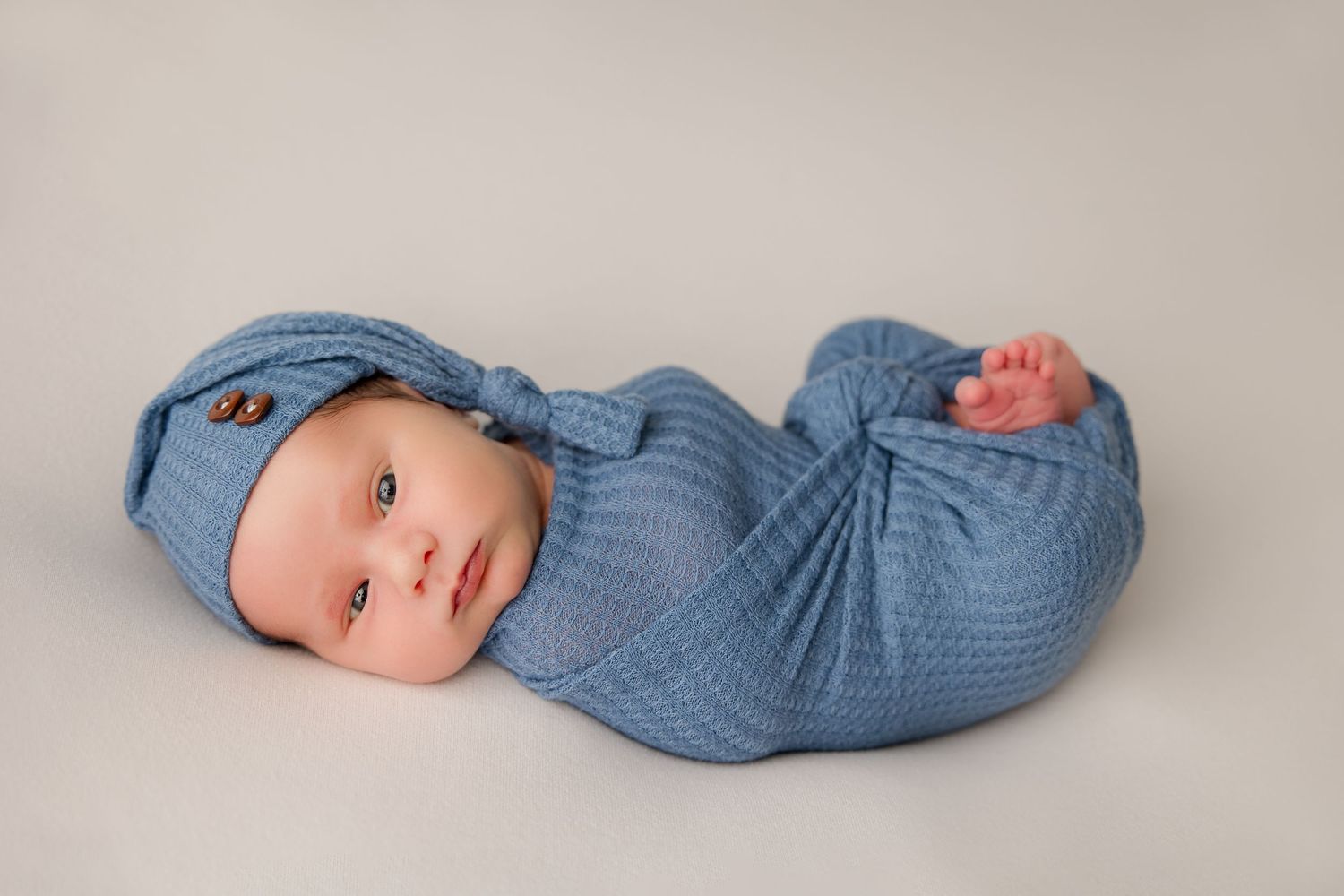 Sleeping newborn baby wearing blue outfit lies peacefully on white backdrop.