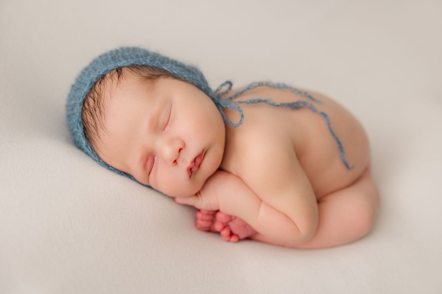 Sleeping newborn wearing blue knit hat lying on white backdrop in peaceful pose.