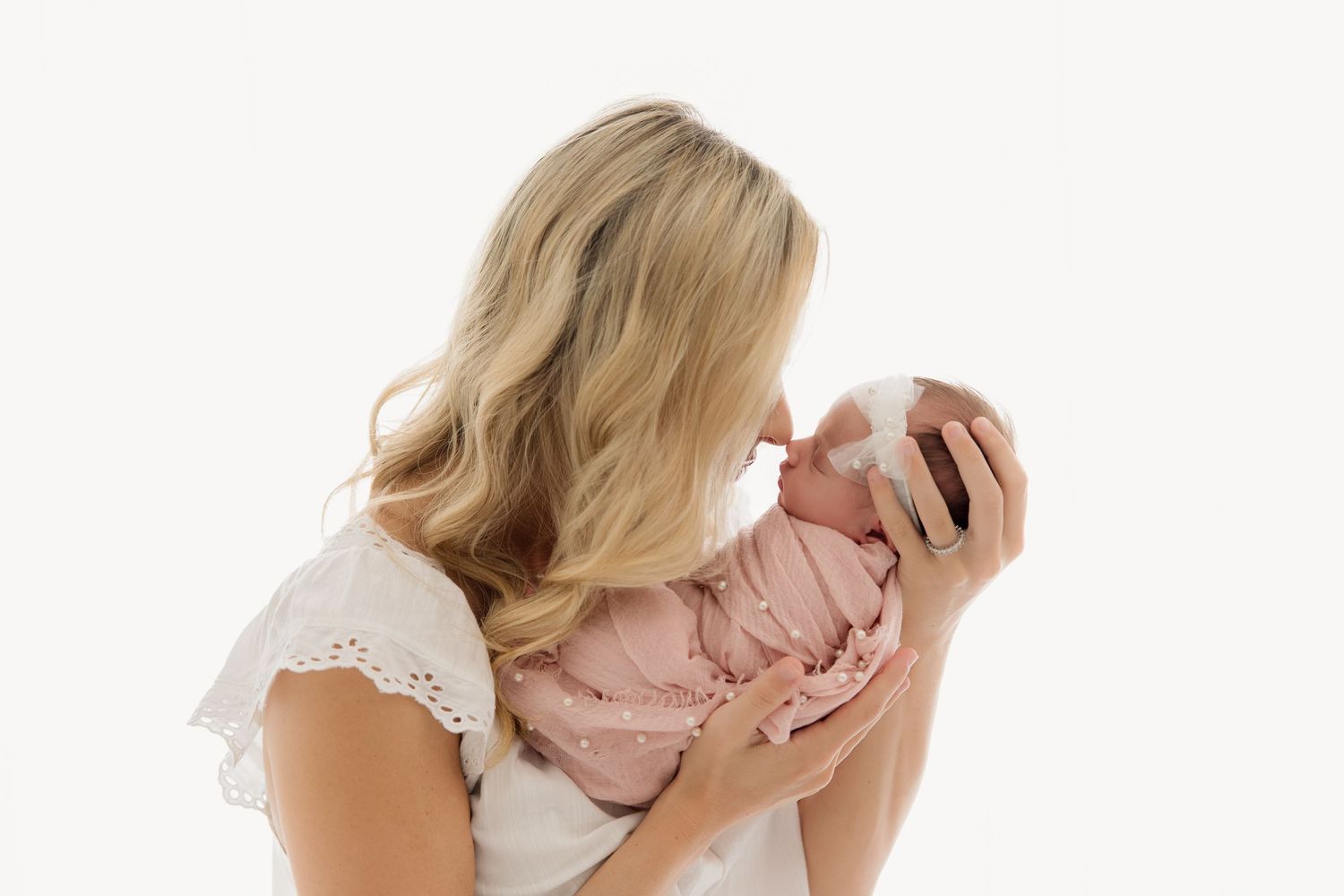 Parent in white lace dress holds newborn baby swaddled in pink against white background.