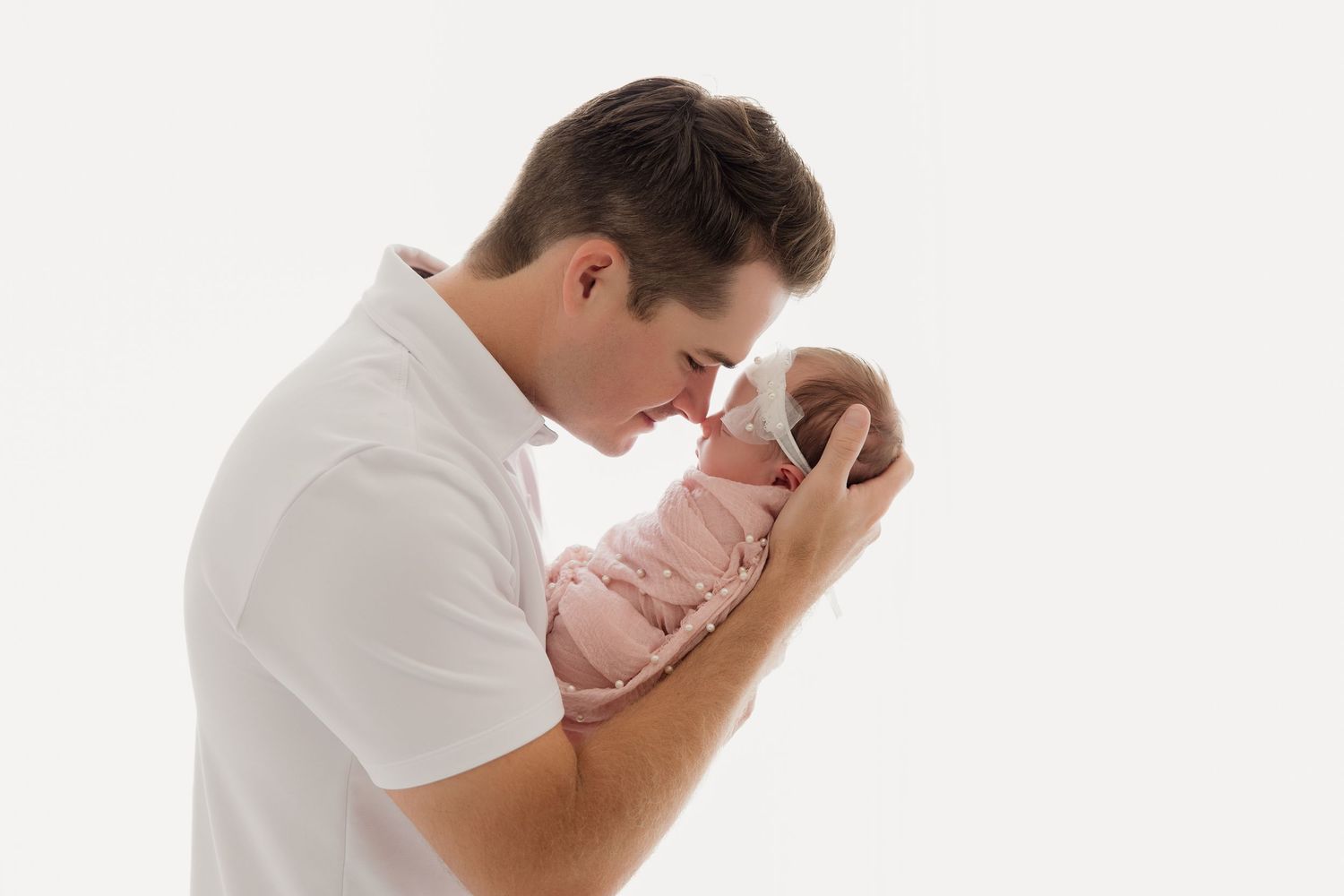 Parent in white shirt tenderly embraces sleeping newborn wrapped in pink against white backdrop.