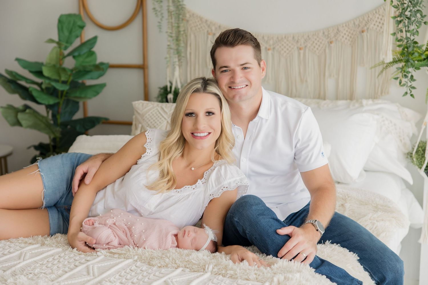 A couple shares affectionate moments while sitting on a bed with greenery and decor in the background.