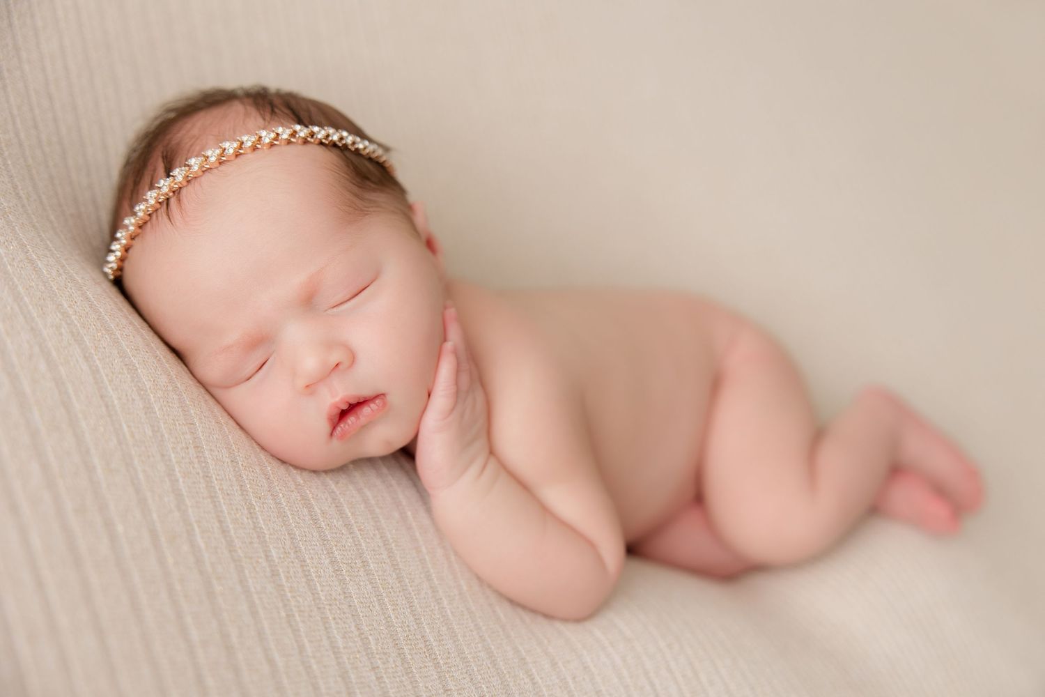 Newborn baby wearing delicate headband sleeping peacefully on soft beige fabric.