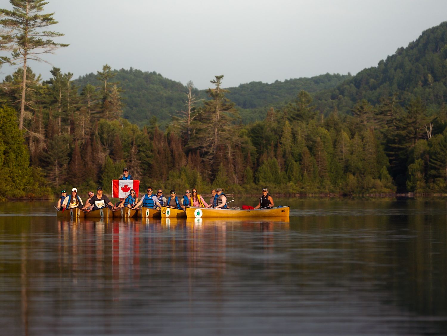 Canoes with Canadian flags glide across a serene lake surrounded by evergreen trees and mountains at sunset.
