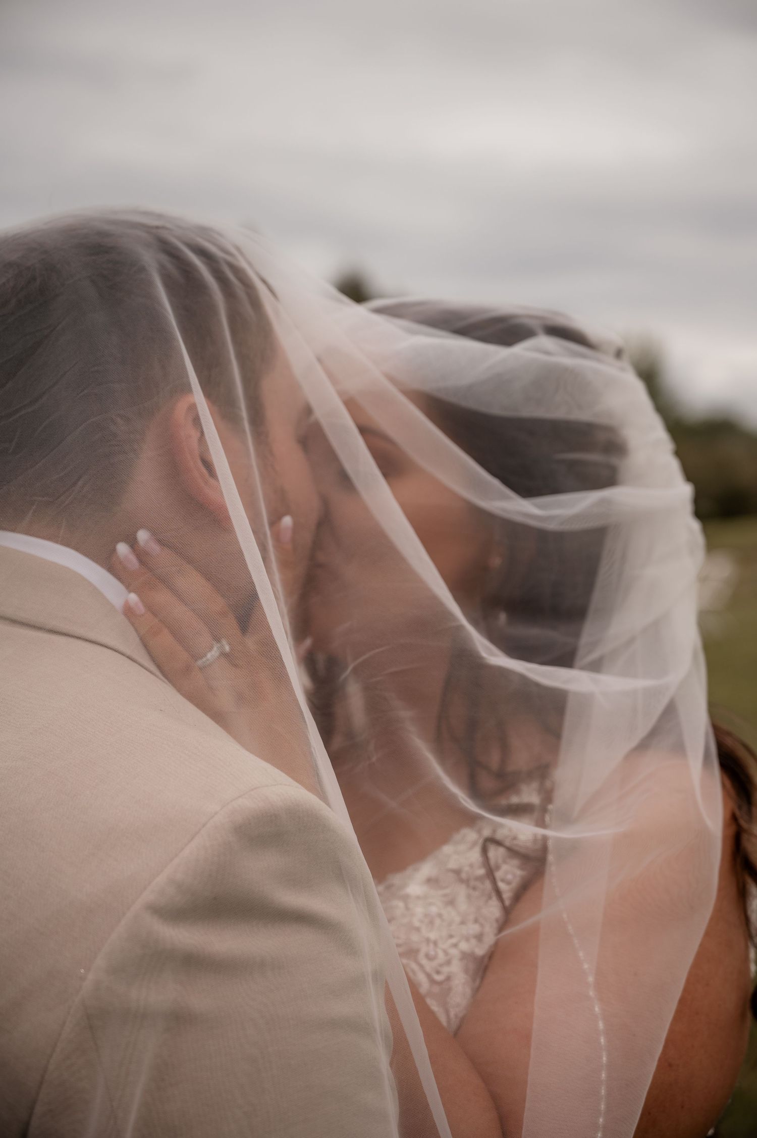 Bride and groom sharing a romantic kiss under a flowing white wedding veil outdoors.