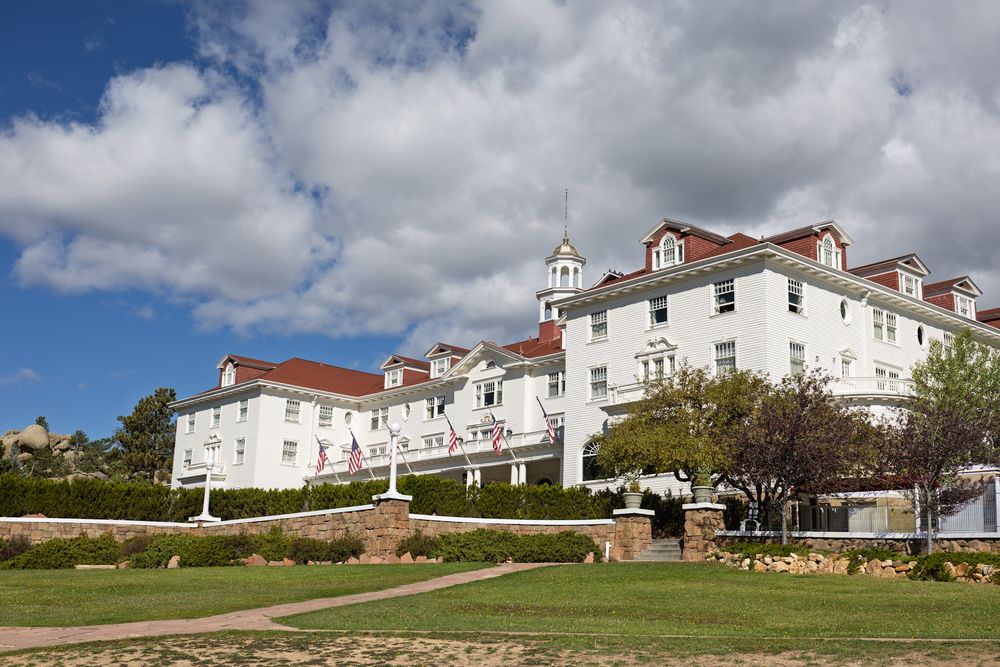 Grand white Stanley Hotel building with red roof sits majestically on a landscaped hillside under blue skies.