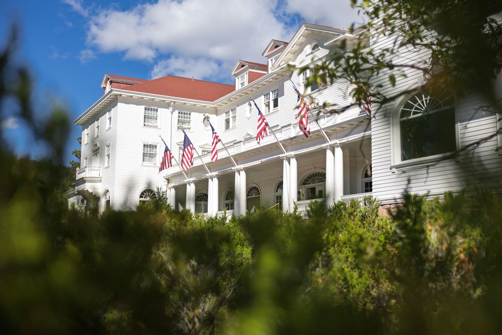 Historic Stanley Hotel with white columns and American flags viewed through foliage on a sunny day.