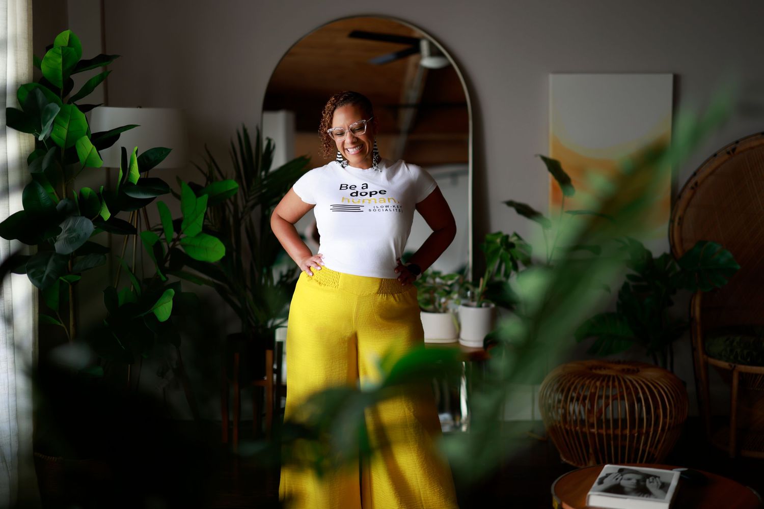 A person stands in a bright interior space wearing a white t-shirt and yellow pants amidst lush green houseplants.