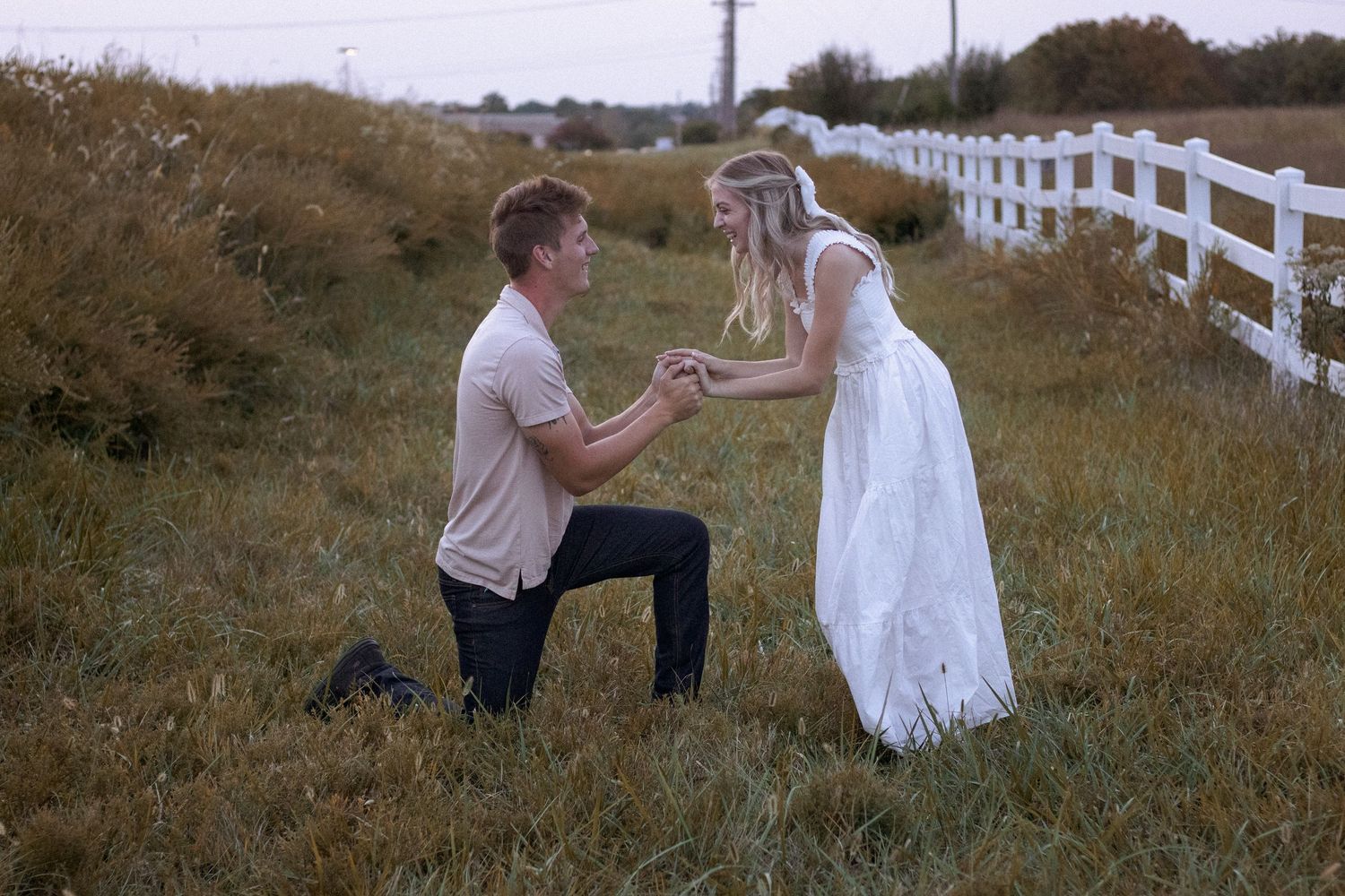 Couple in white dress and casual wear on grassy hill with white fence.