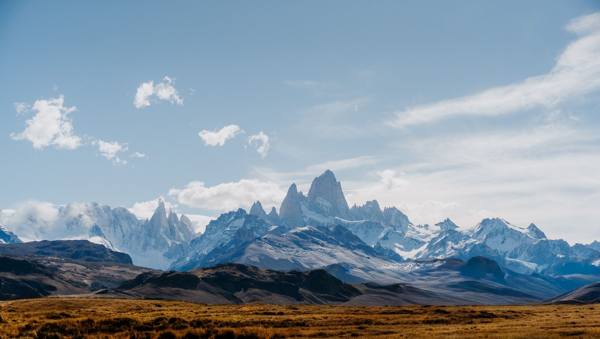 A panoramic series of mountain peaks with snow-capped summits against a blue sky with white clouds in Patagonia.