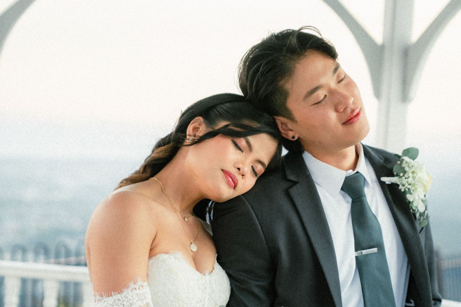 Bride in an off-shoulder white gown rests her head on the groom's shoulder in a dark suit during a romantic rooftop wedding portrait.