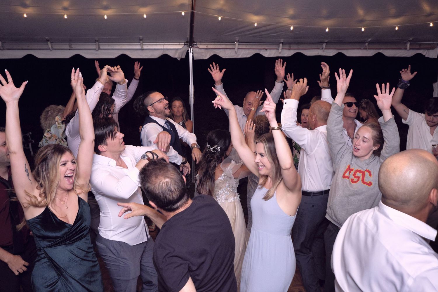 People dancing together under a tent decorated with sparkling string lights at an outdoor wedding reception.