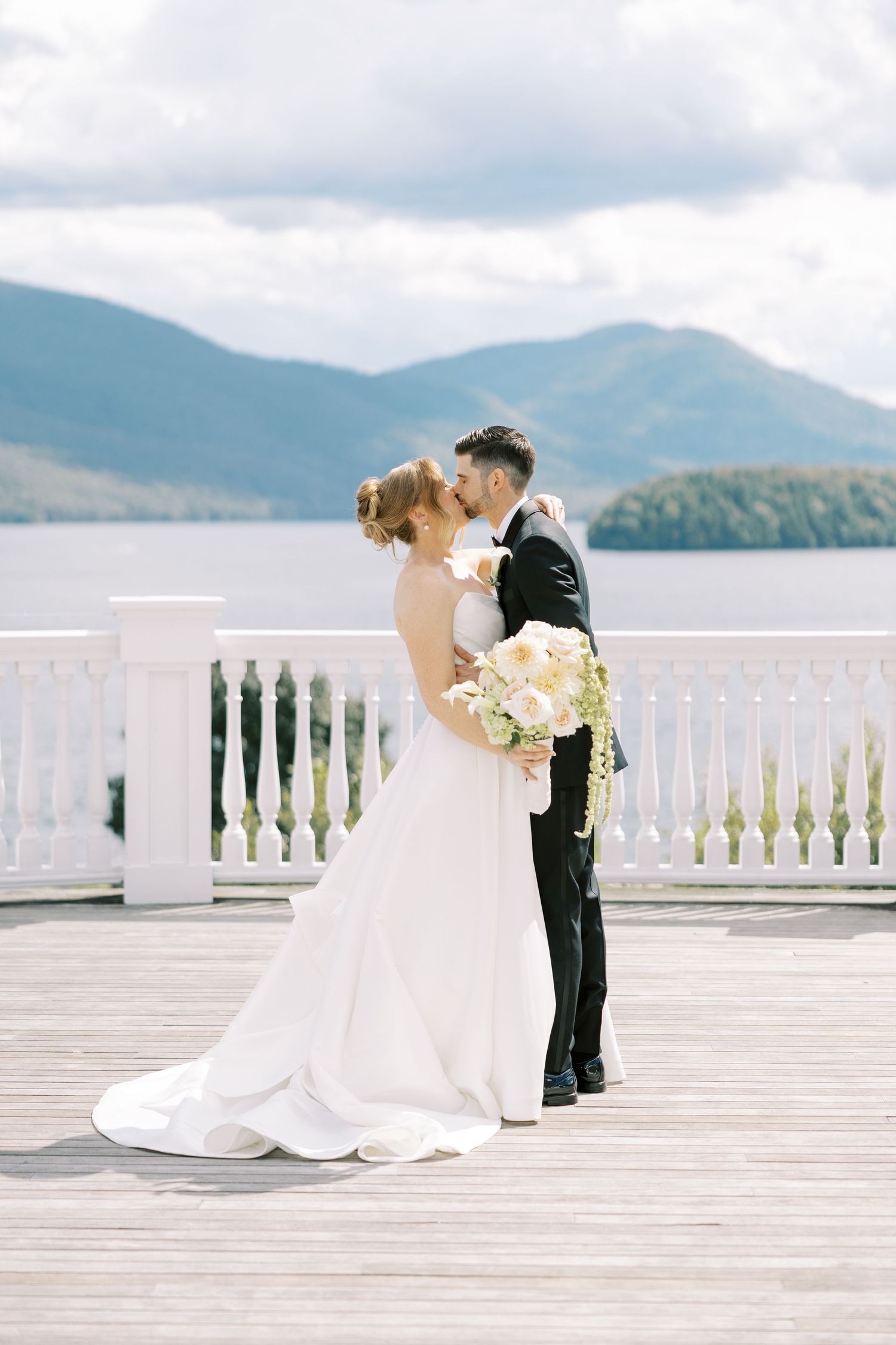 Elegant couple in wedding attire stands on a waterfront terrace with white railings overlooking a scenic mountain lake view.