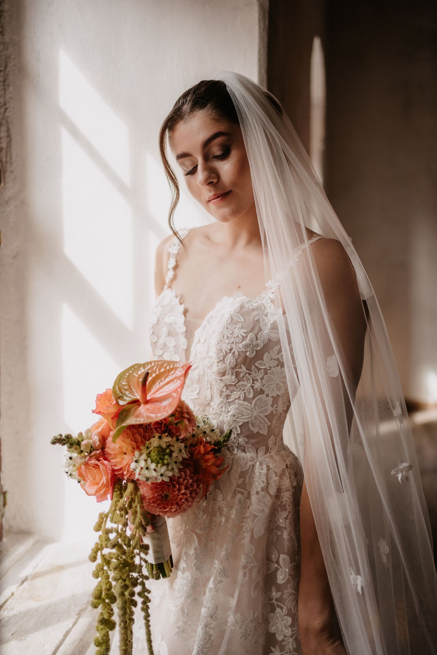Elegant bride in lace wedding dress and veil poses near window with dramatic lighting and autumn-colored floral bouquet.