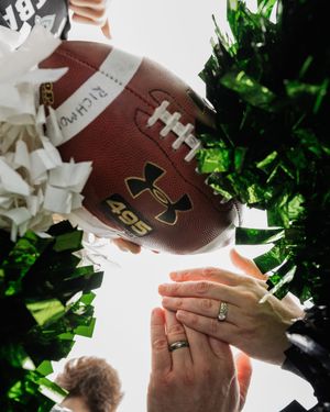 A couple holds a football together while showing their wedding rings in a creative family photo.