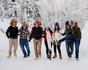 Family of 7 jump playfully in the snow wearing coordinated winter outfits of khaki pants and dark tops.