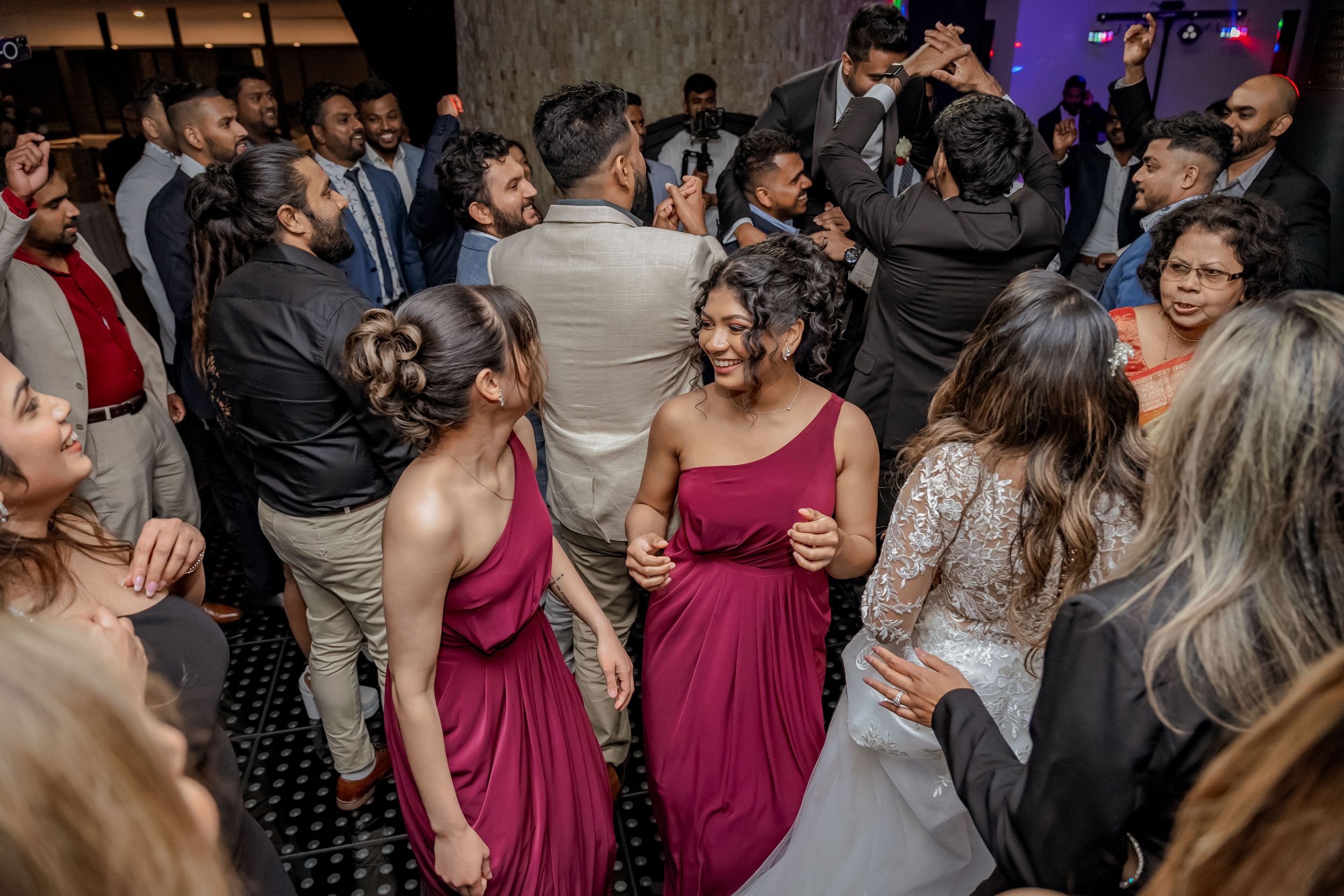 Wedding guests dance and celebrate on a crowded dance floor at an evening reception while wearing formal attire.
