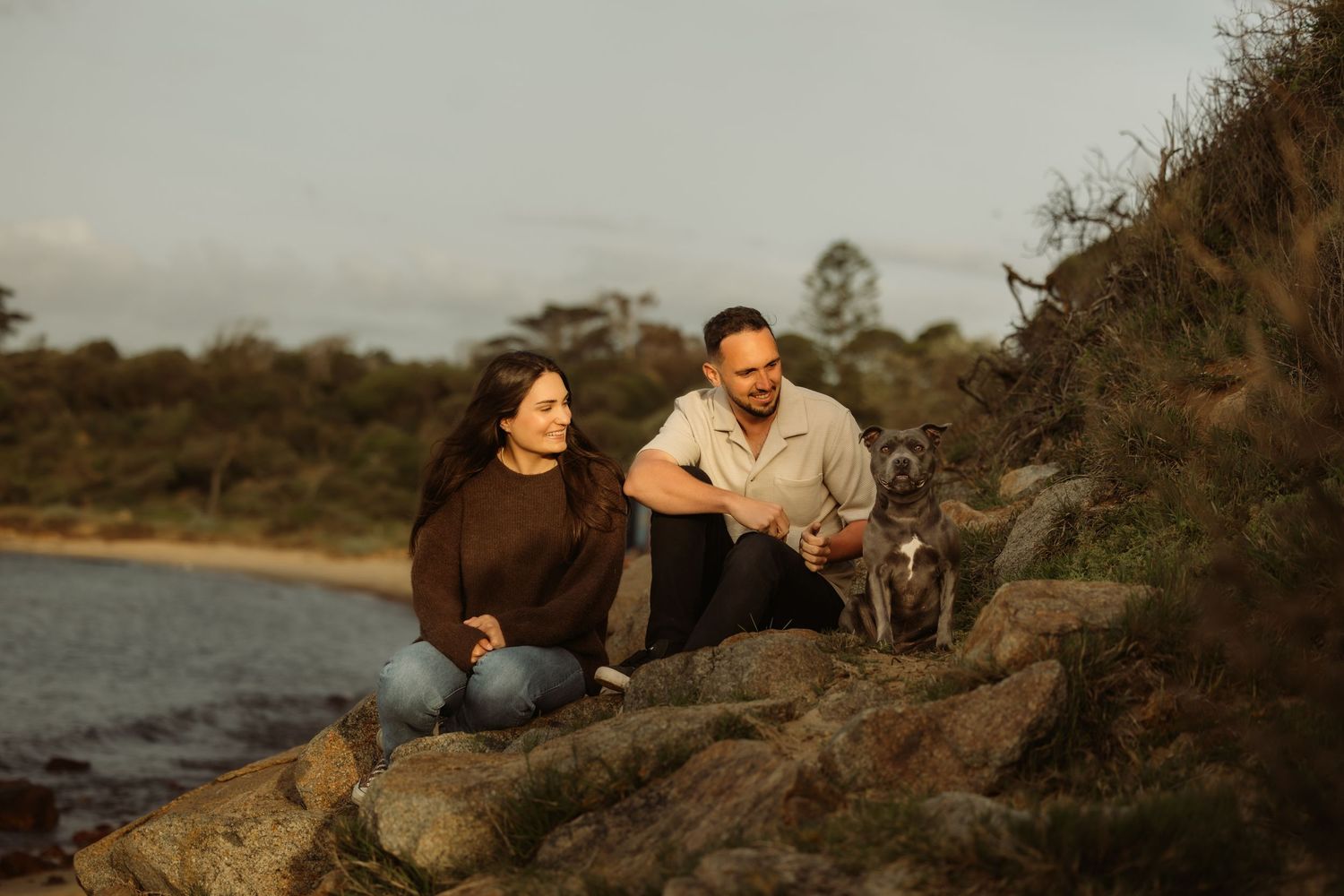 Couple sitting on rocky shoreline with their blue staffy dog, surrounded by coastal bushland at golden hour.
