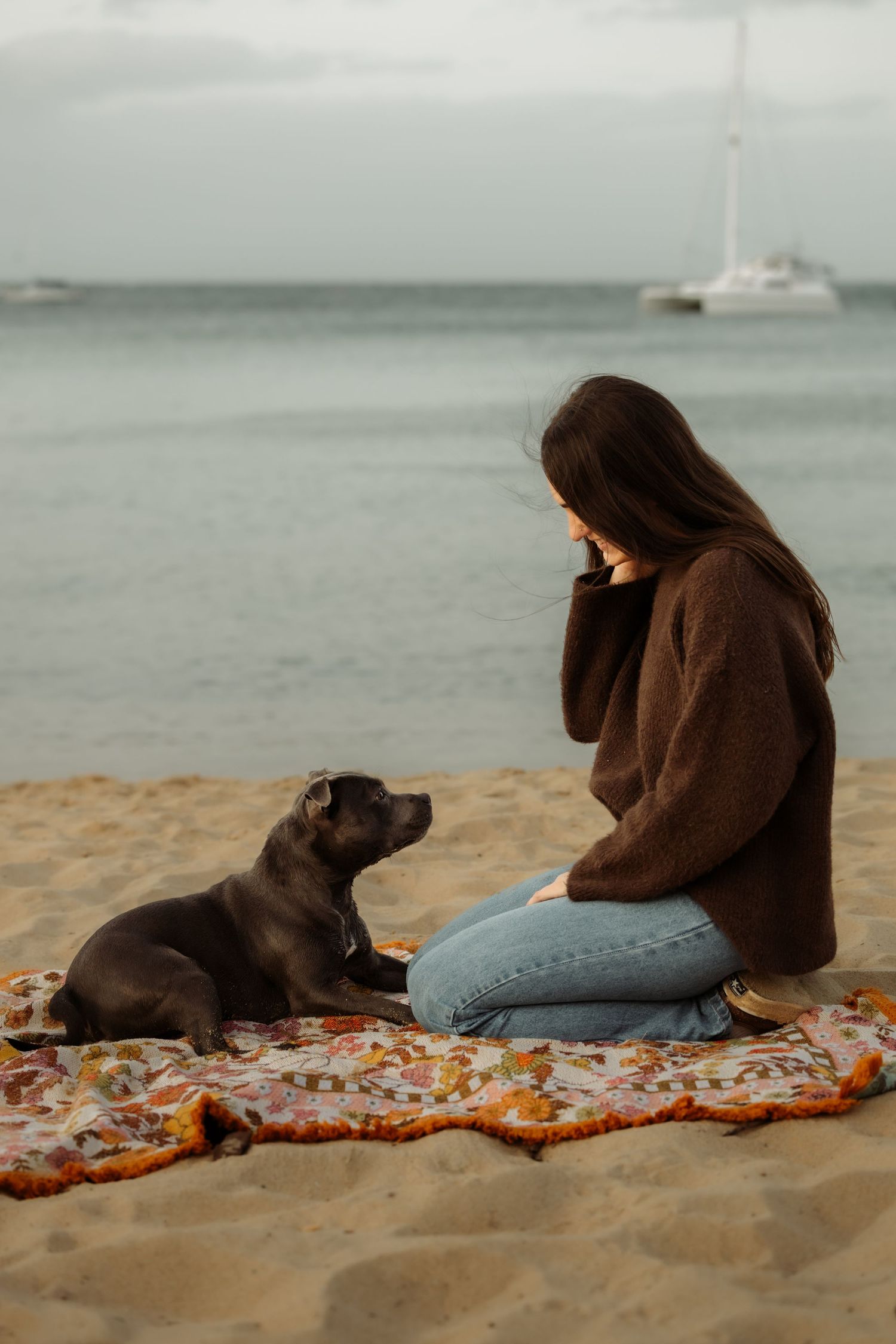 A young woman in a brown sweater and jeans sits on a floral blanket on the beach with her black dog.