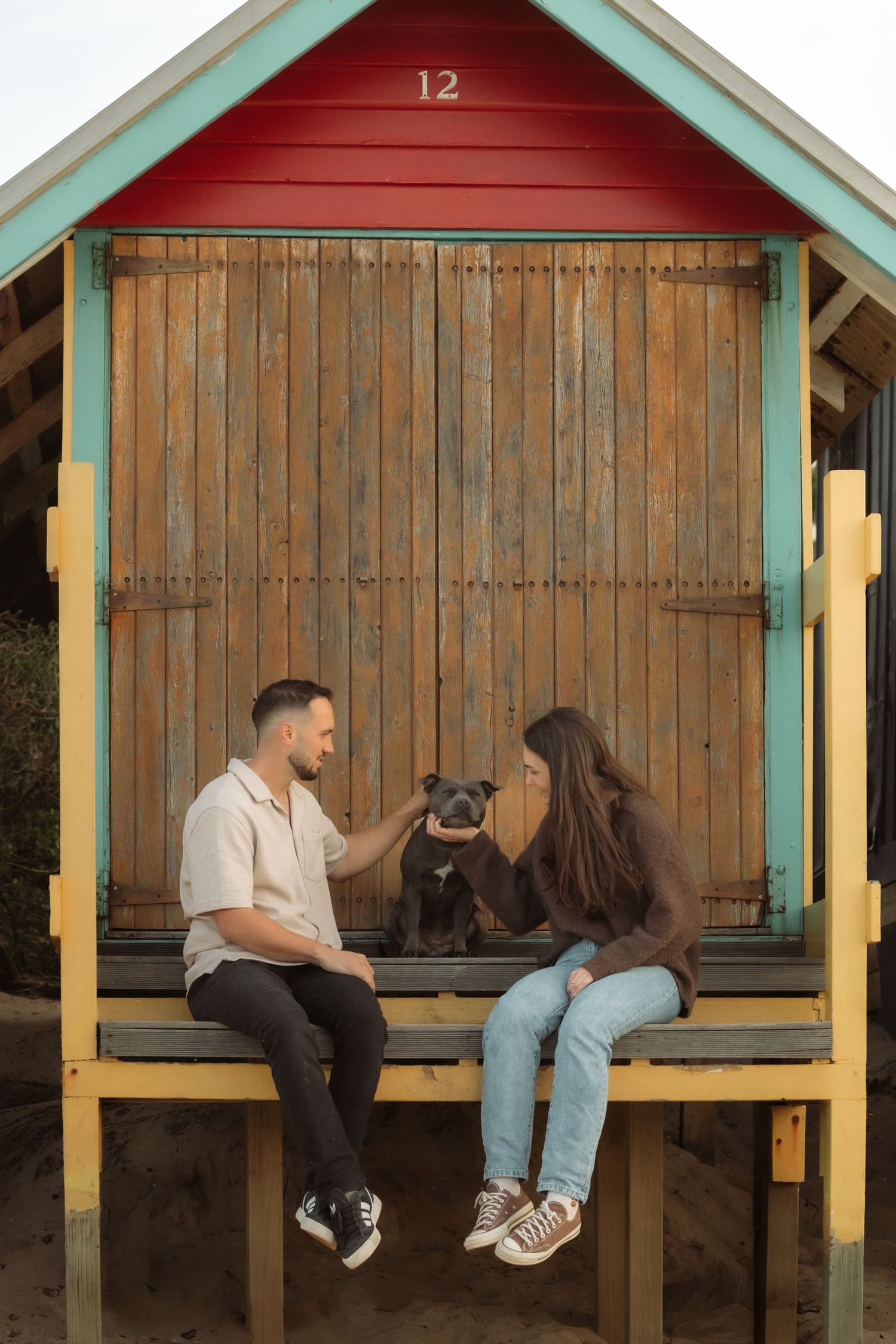 Couple sitting with black dog at colorful beach hut number 12 with wooden shutters.