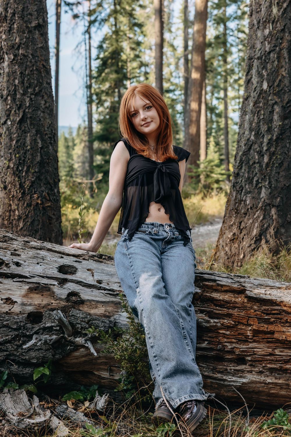 Woman in casual attire poses on a fallen log in a pine forest setting.