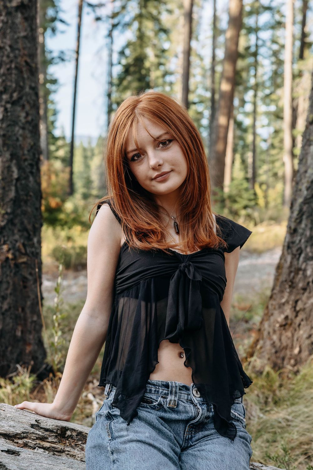 Portrait of a young woman in black top and jeans sitting on a fallen log in a forest.