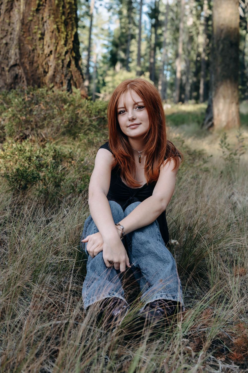 Portrait of a woman sitting in tall grass surrounded by evergreen trees.