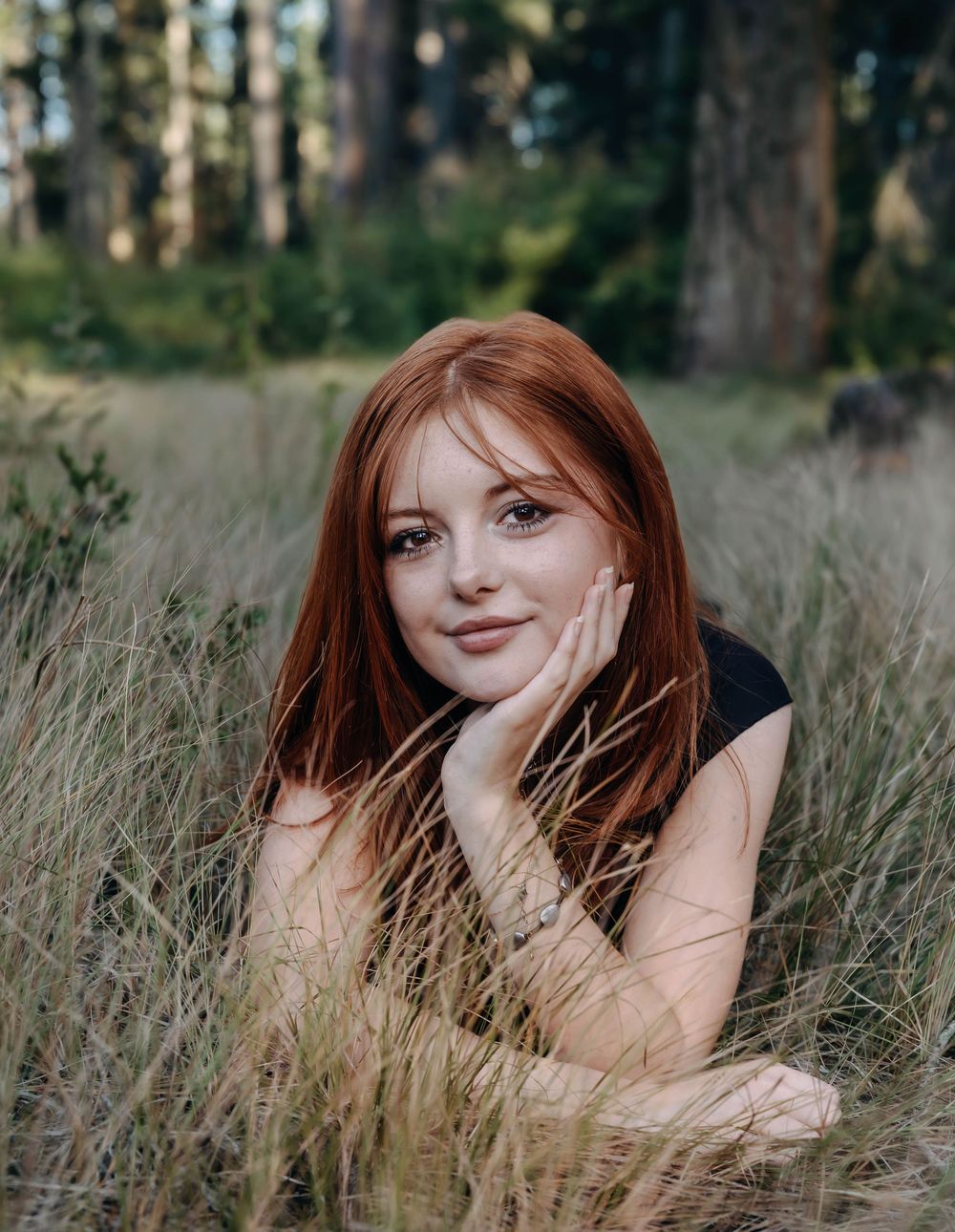 Woman with auburn hair lying in tall grass with forest backdrop.