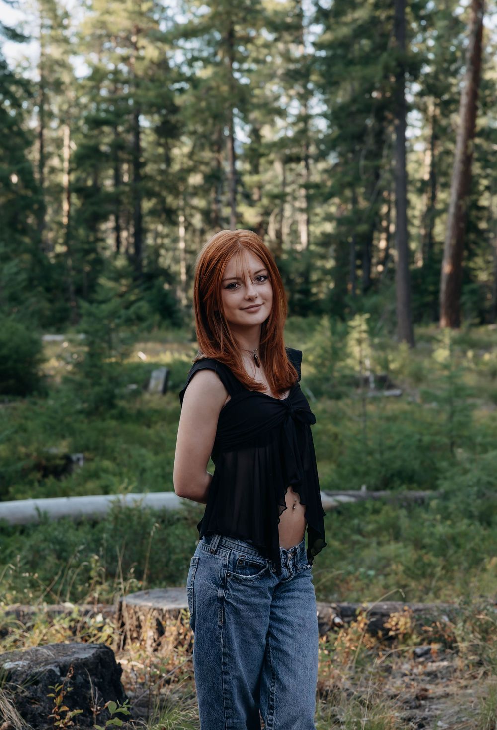 Woman in black top stands against pine forest background during golden hour.