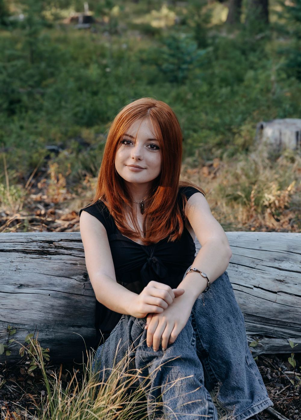 A person wearing jeans sits casually on a fallen log in a wooded outdoor setting.