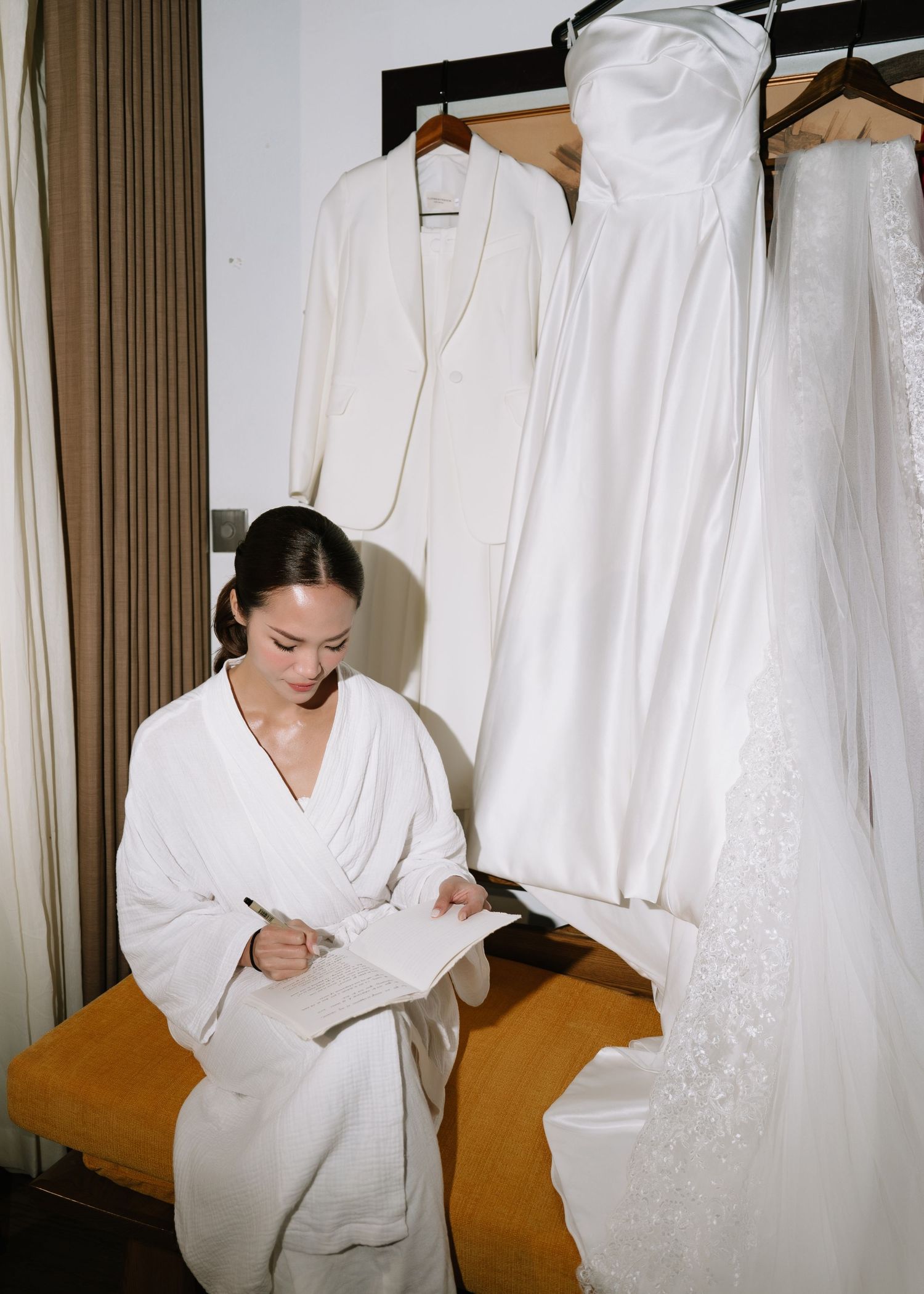 Bride in white robe writing notes beside hanging wedding gown and white suit in bridal suite.