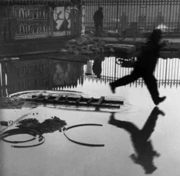 Black and white photograph of a silhouetted figure leaping over a puddle, casting a reflection in the wet pavement.