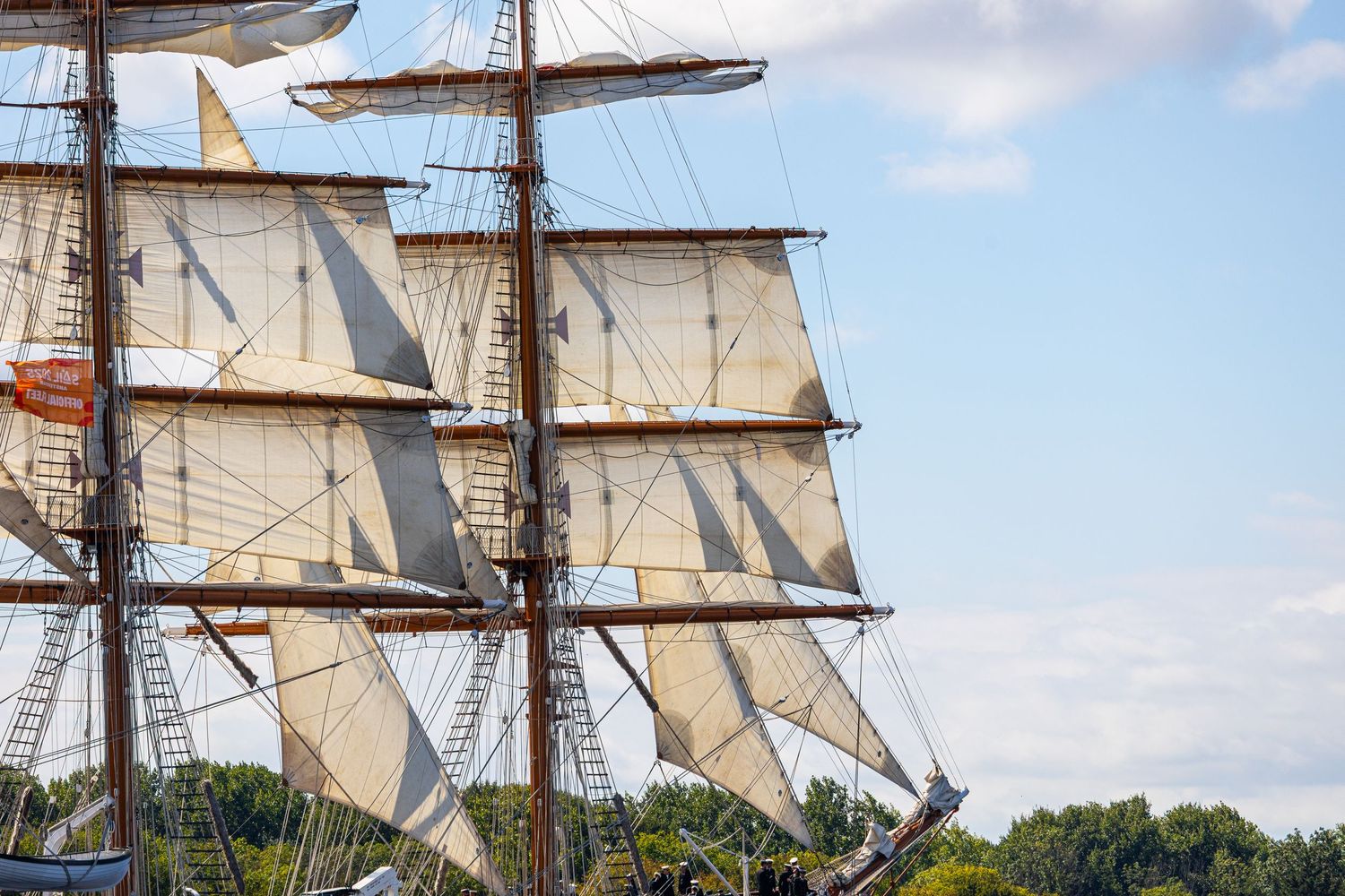 White canvas sails and tall masts of a historic sailing ship against a blue sky with trees in the background.