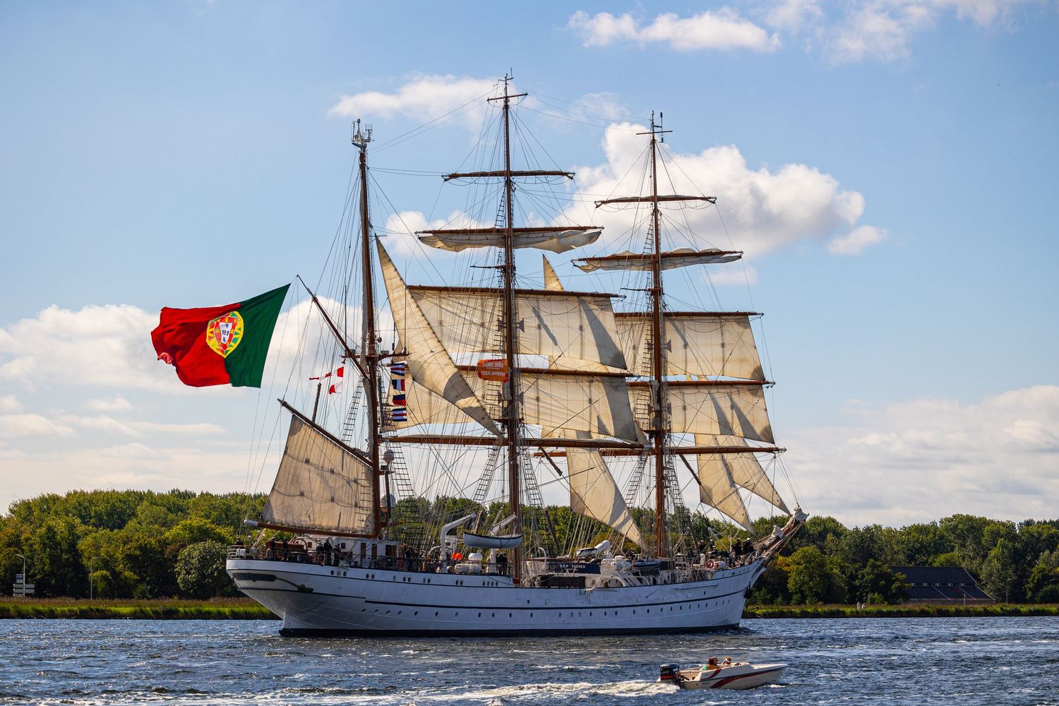 A tall ship sailing vessel with white sails glides across calm waters under a blue sky with puffy clouds.