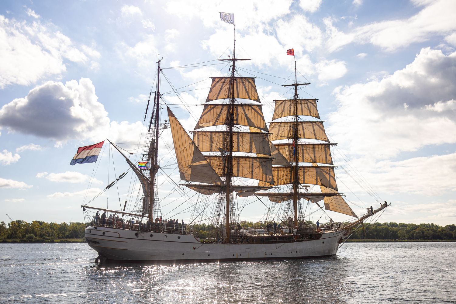 A series of tall ships with white sails glide across the water under a cloudy sky during a maritime festival.
