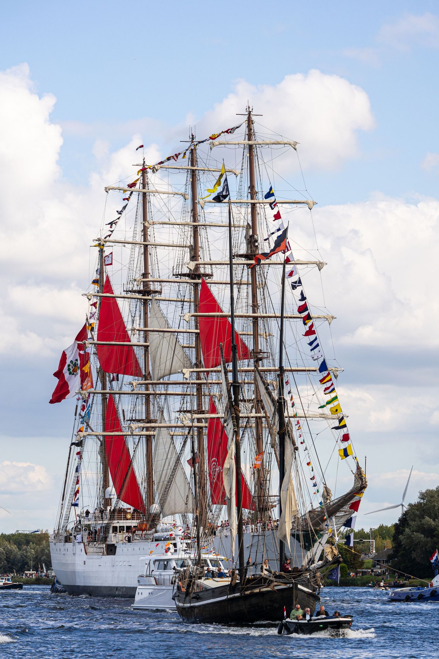Tall ships with colorful signal flags and red sails sailing in formation during a maritime festival on blue waters.