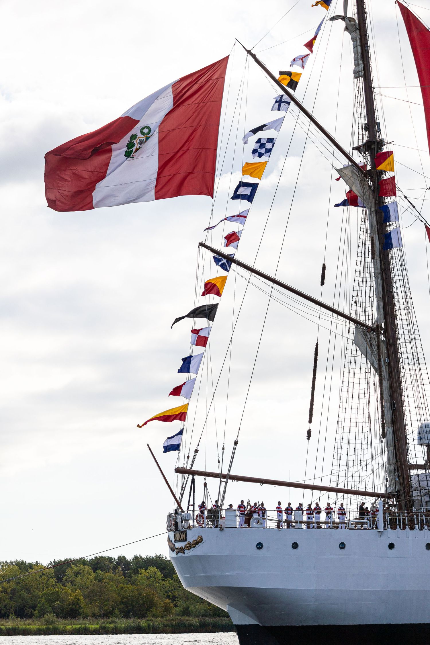 Tall ship with colorful nautical flags flying from its masts sails across calm waters under cloudy skies.