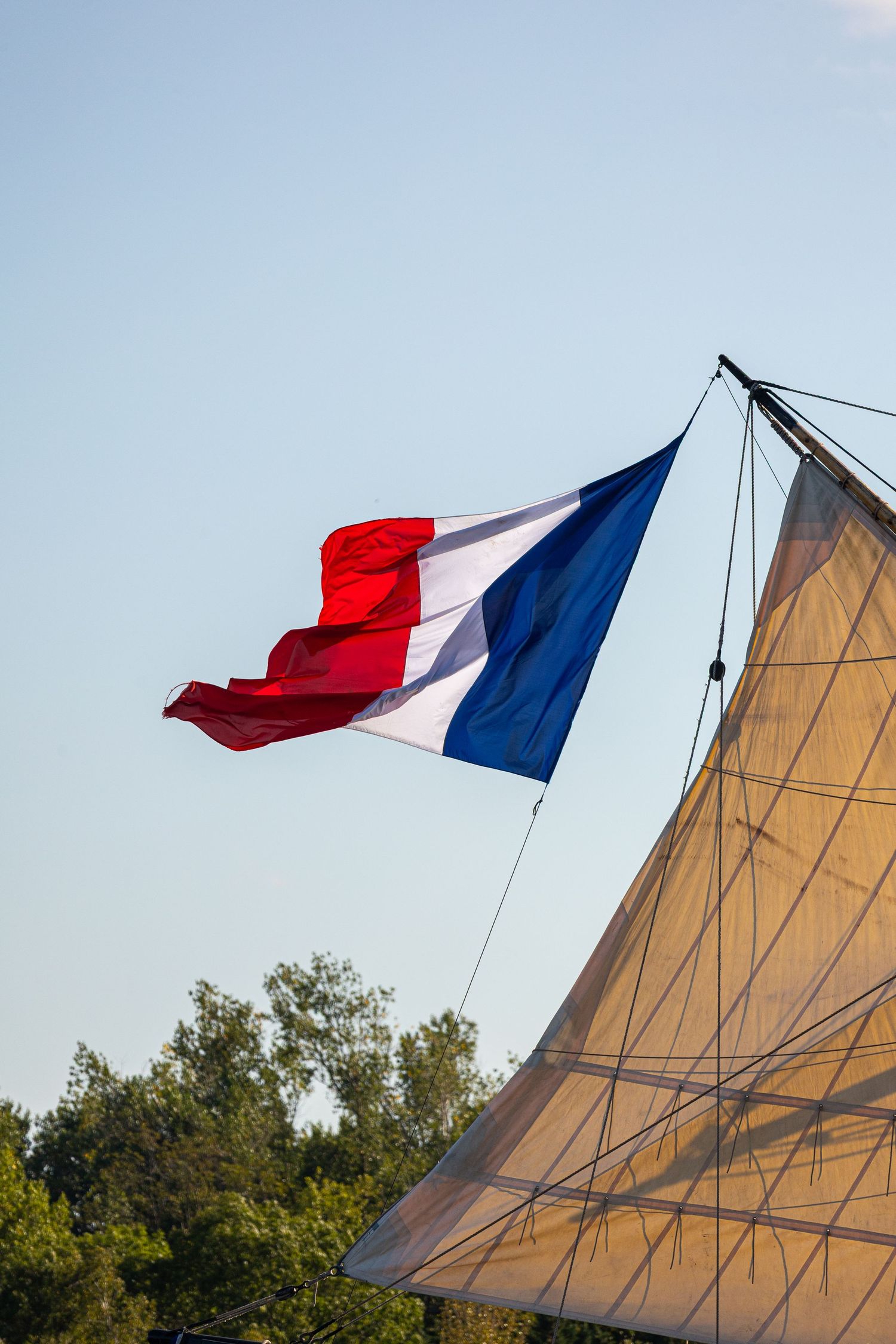 French flag waves proudly against blue sky above tan-colored sailboat canvas in summer scene.
