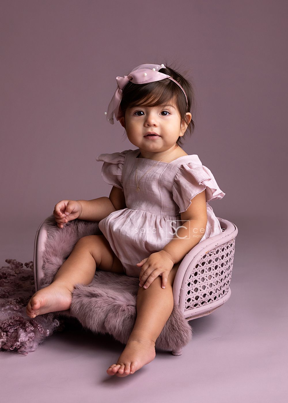 Baby in pink dress sits on pink cushion with plush toy in studio setting.