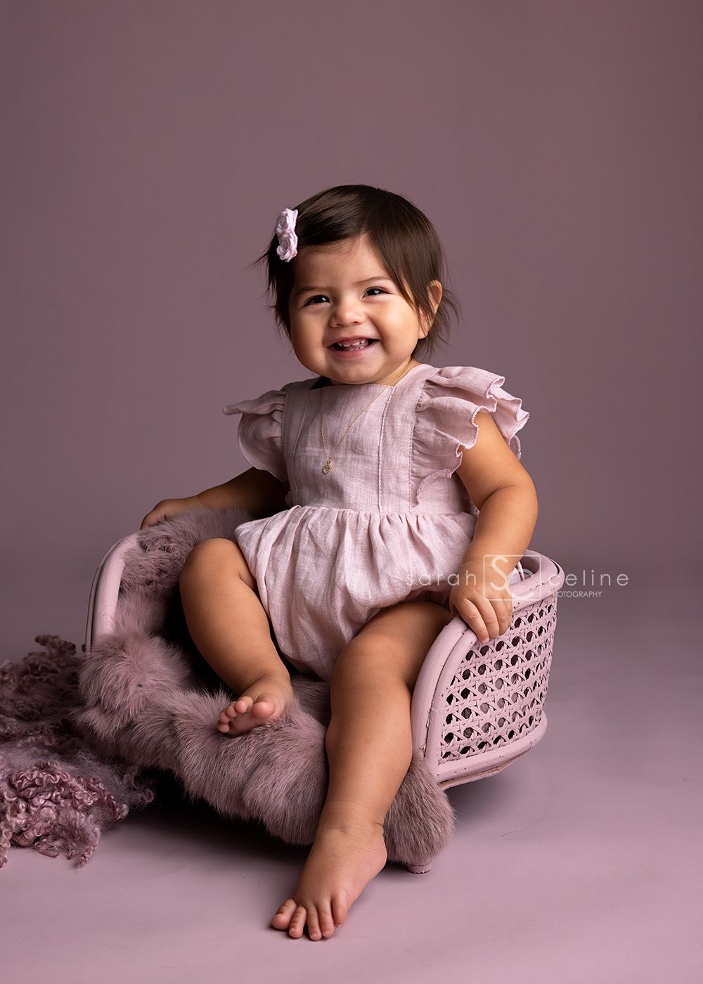 Baby wearing ruffled pink dress poses on pink cushion with soft lighting.